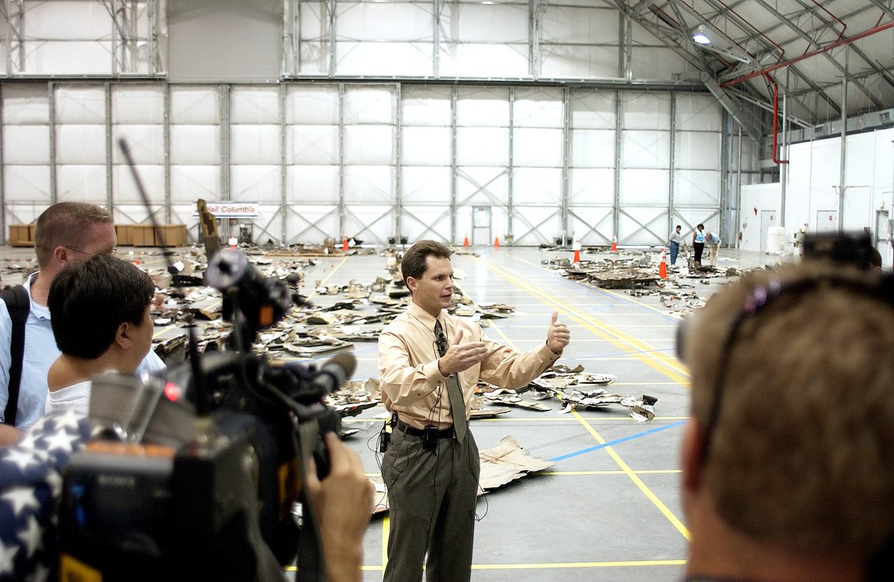 Scott Thurston discusses Columbia debris storage at Kennedy Space Center, surrounded by packed debris in hangar.