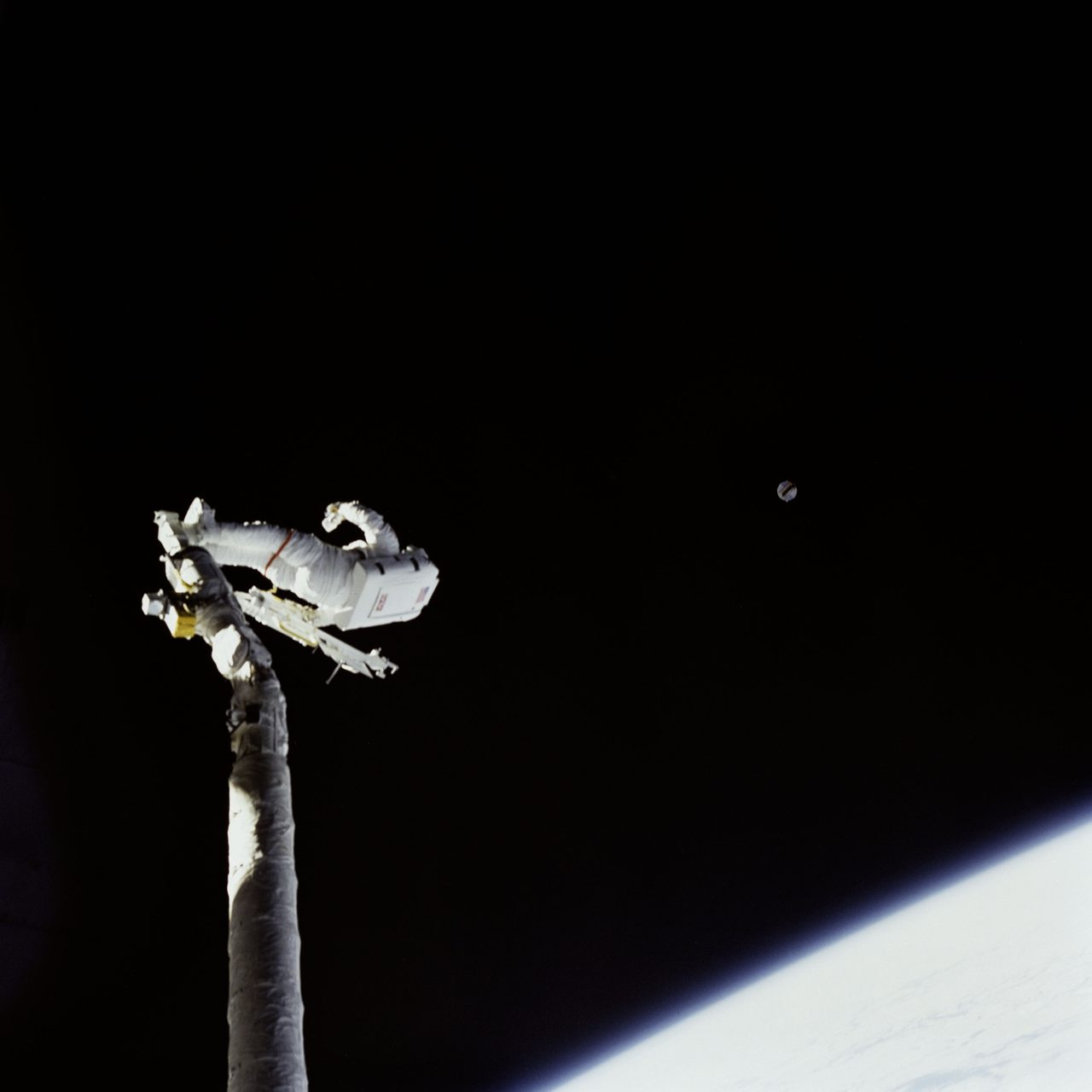 Astronaut James van Hoften operates robotic arm tracking satellite Syncom IV-3 in space.
