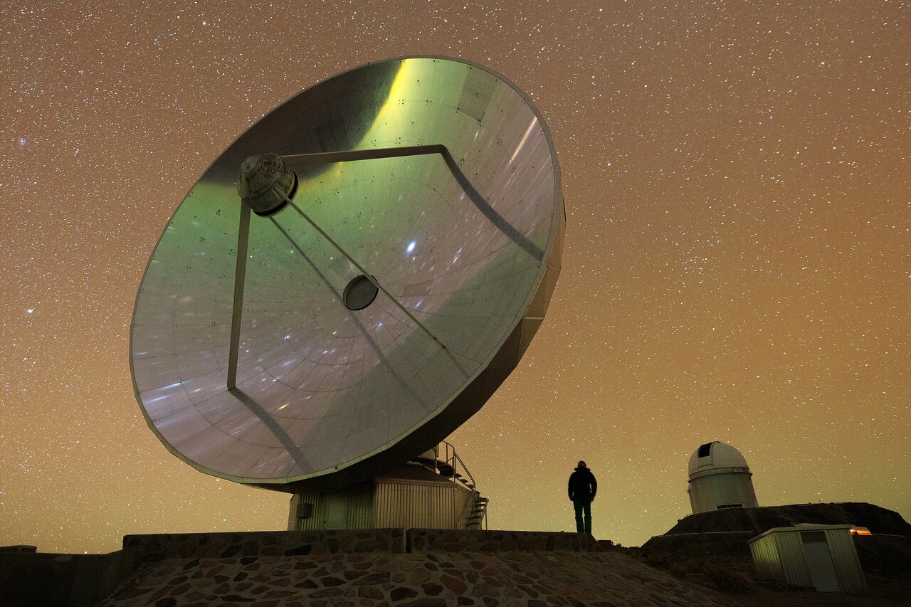 Large satellite dish reflecting a clear blue sky, surrounded by green hills and scattered white clouds.