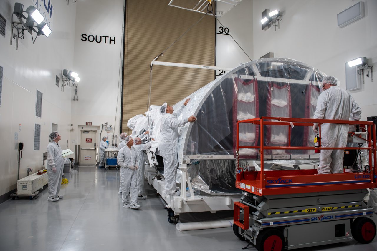 A large Atlas V rocket fairing being transported on a flatbed truck under clear skies.