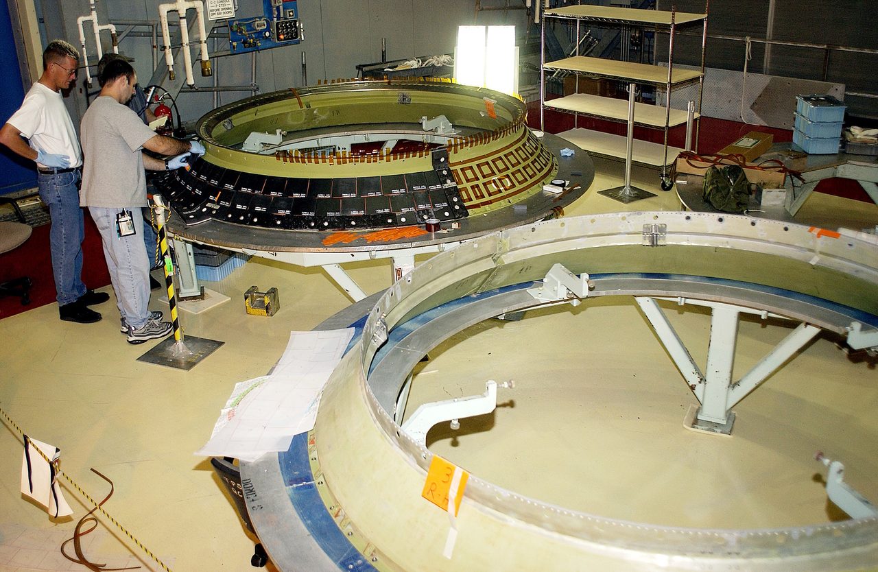 Three engineers at Kennedy Space Center install protective heat shield tiles on the Discovery orbiter's main engine.