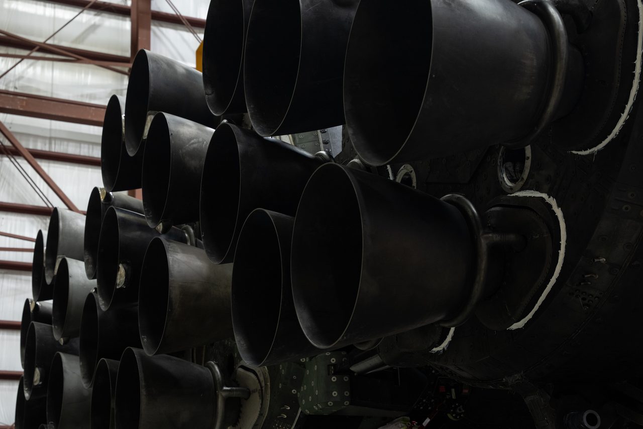 A yellow crane lifts SpaceX Falcon Heavy rocket with 27 engines inside an industrial facility for NASA's Roman Space Telescope.