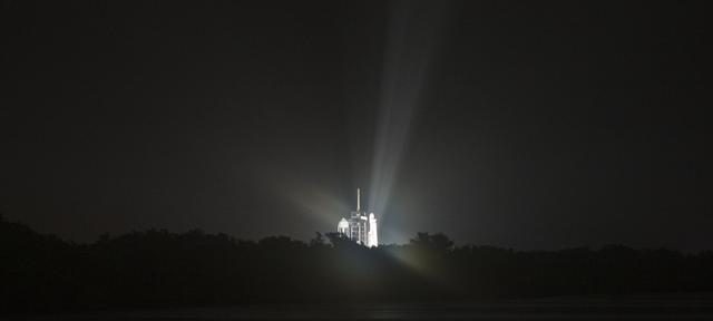 SpaceX Falcon Heavy rocket launching at night with fiery exhaust and smoke against a starry sky.