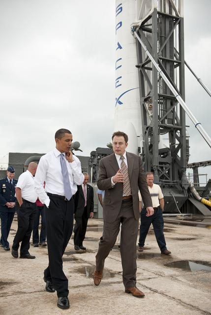 President Obama touring Kennedy Space Center, surrounded by NASA officials and space shuttle models.