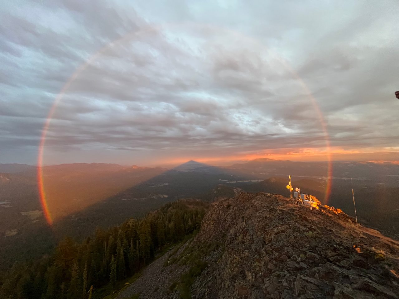 A vibrant rainbow arches over a shadowy landscape with dark clouds and a hint of sunlight breaking through.