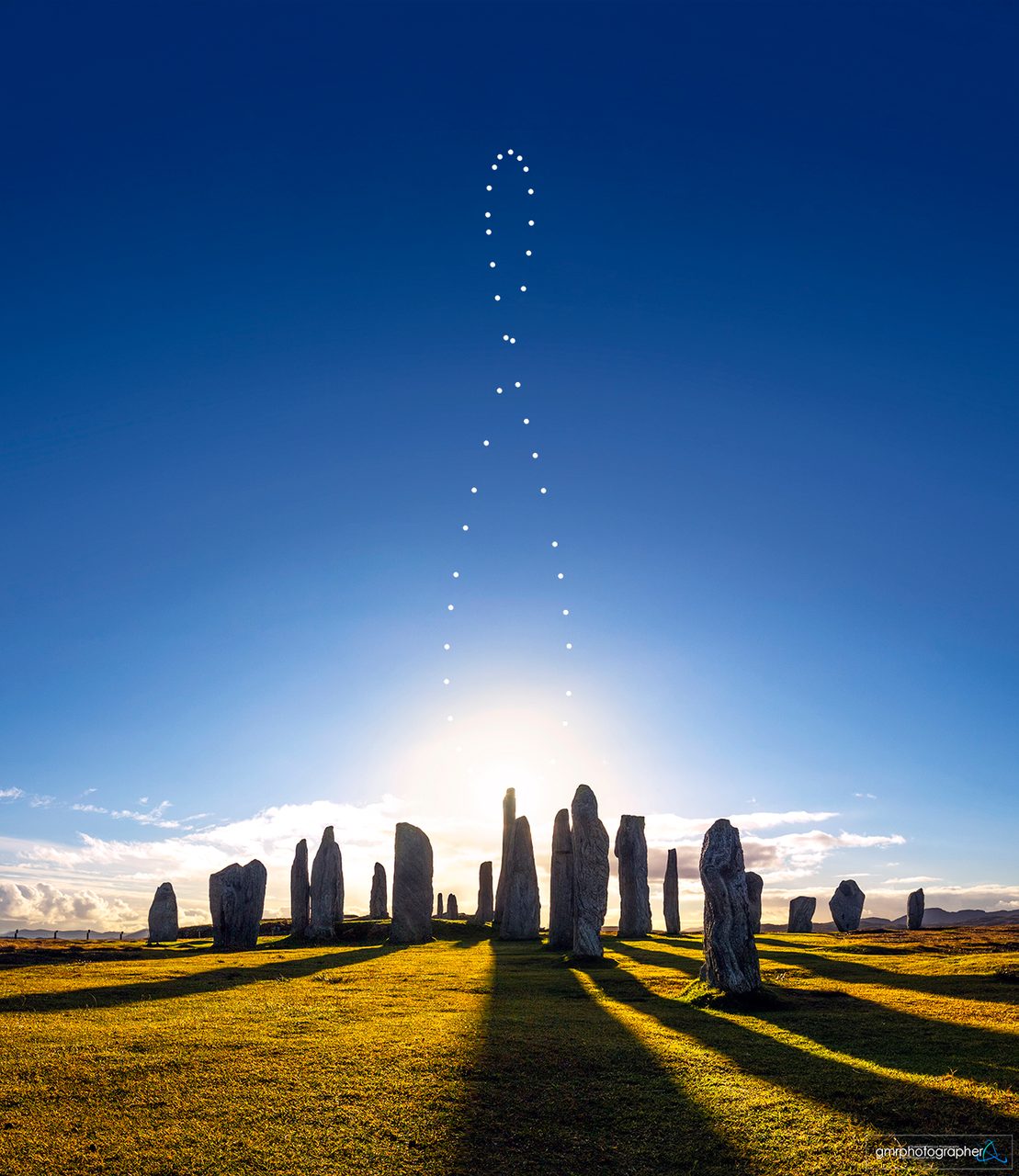 A celestial analemma pattern arcs over the ancient Callanish Stones under a clear blue sky.