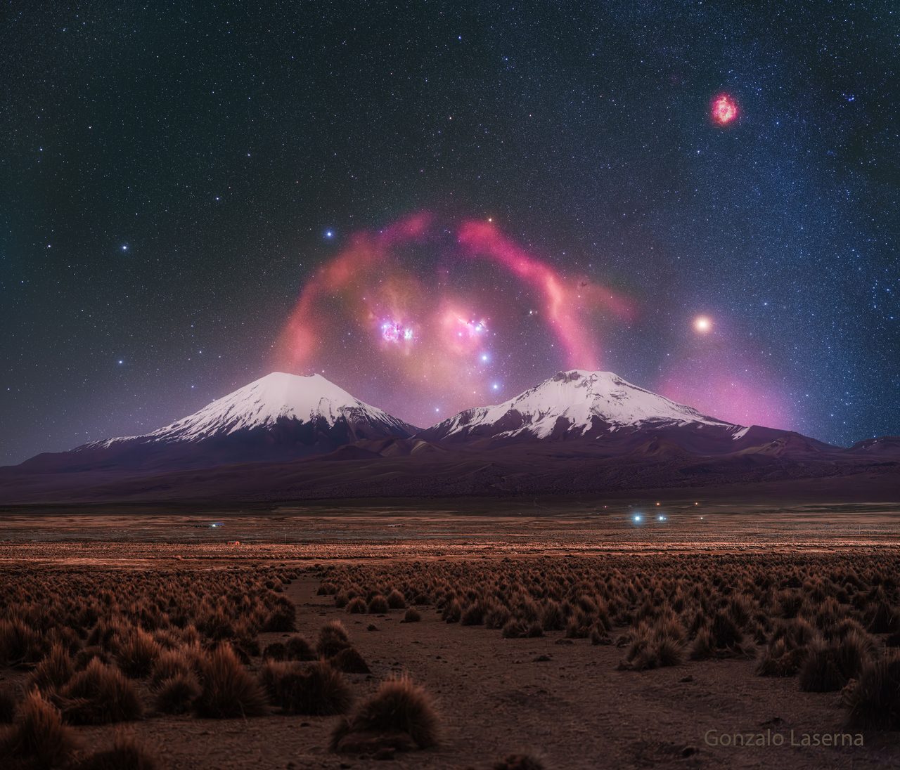 A vibrant arc of Barnard's Loop illuminates the night sky above two silhouetted volcanic peaks.