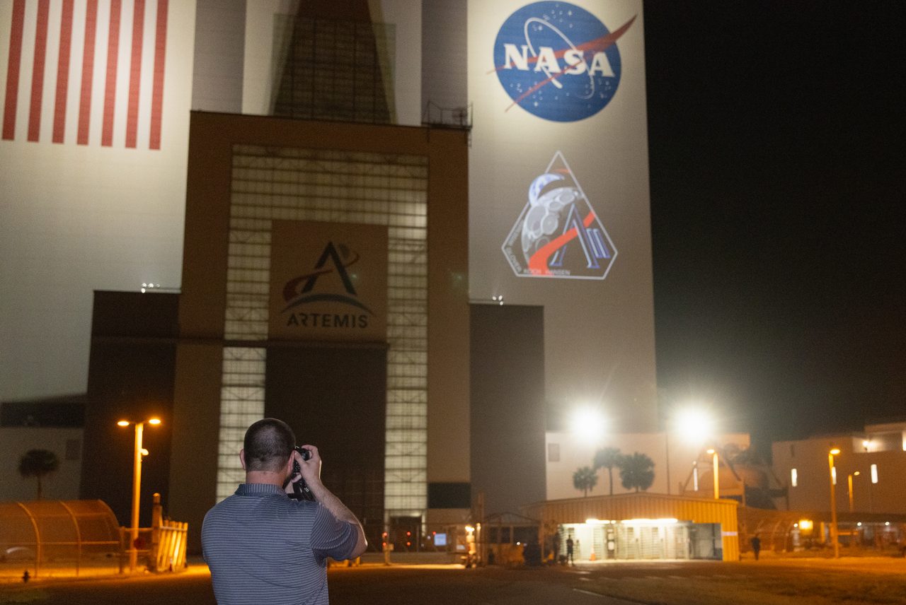 Artemis II mission patch displayed on the Vehicle Assembly Building under a clear blue sky.