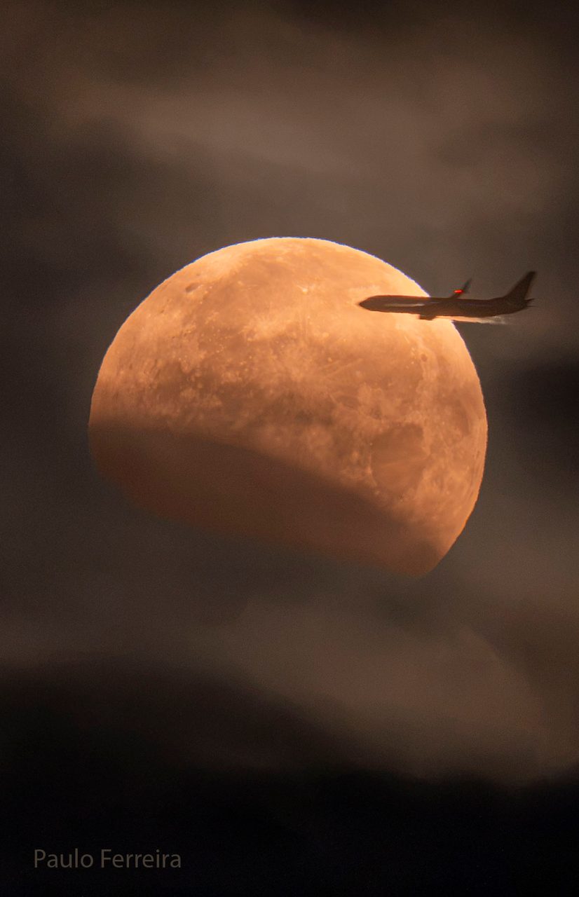 A plane silhouetted against a large, reddish lunar eclipse in a dark night sky.