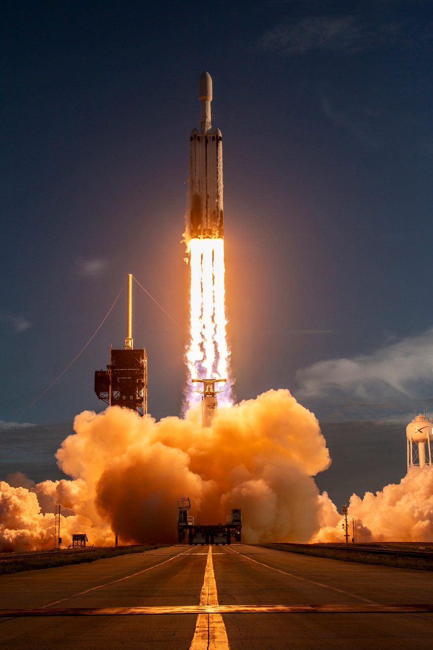 A rocket launches from a coastal pad, emitting bright flames and white smoke against a clear sky.