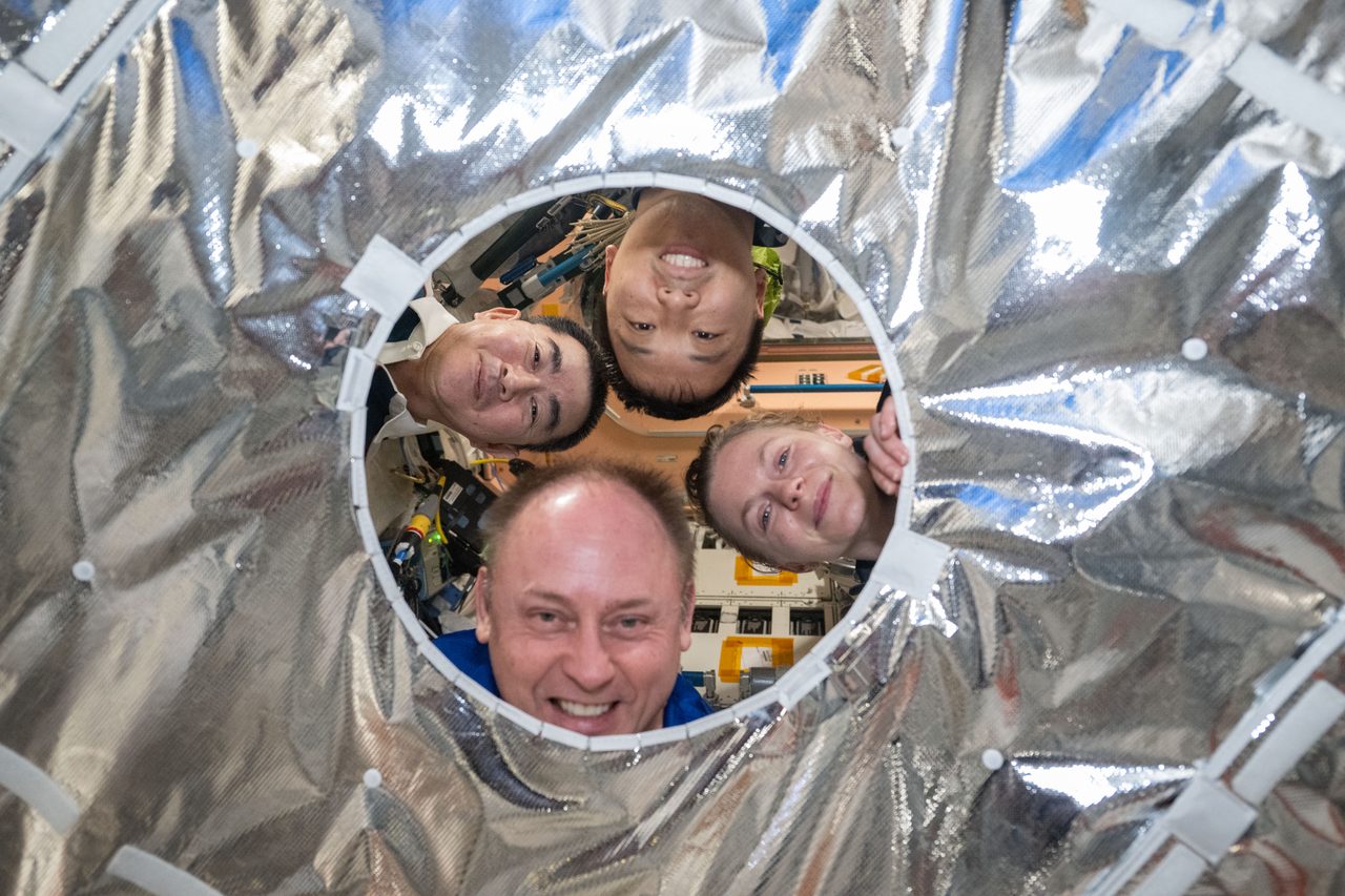 Expedition 73 crew members share a lighthearted moment, peering through a hatch thermal cover aboard the spacecraft.
